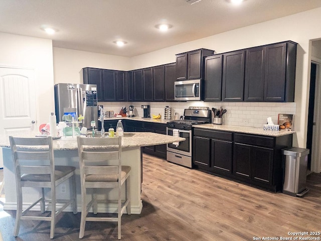 kitchen featuring light wood finished floors, a center island with sink, a breakfast bar area, light stone countertops, and stainless steel appliances