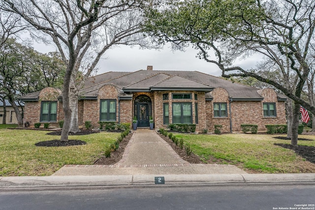 view of front of property featuring a tiled roof, brick siding, a chimney, and a front yard