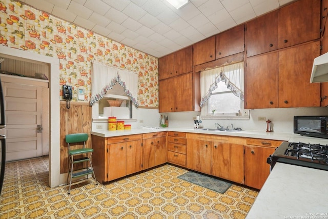 kitchen with wallpapered walls, brown cabinetry, light countertops, black appliances, and a sink