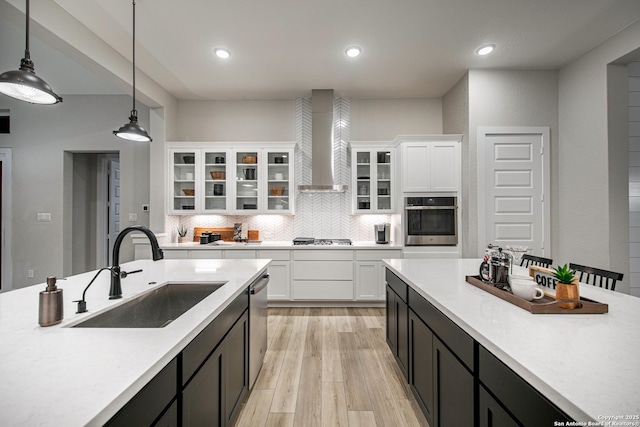 kitchen with a sink, light countertops, wall chimney range hood, hanging light fixtures, and appliances with stainless steel finishes