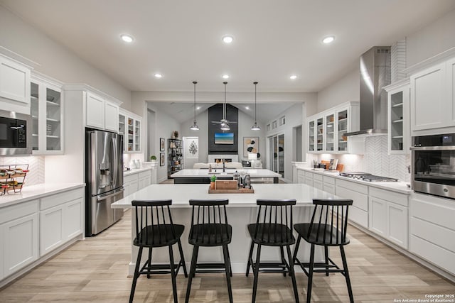 kitchen with open floor plan, light countertops, appliances with stainless steel finishes, wall chimney range hood, and glass insert cabinets