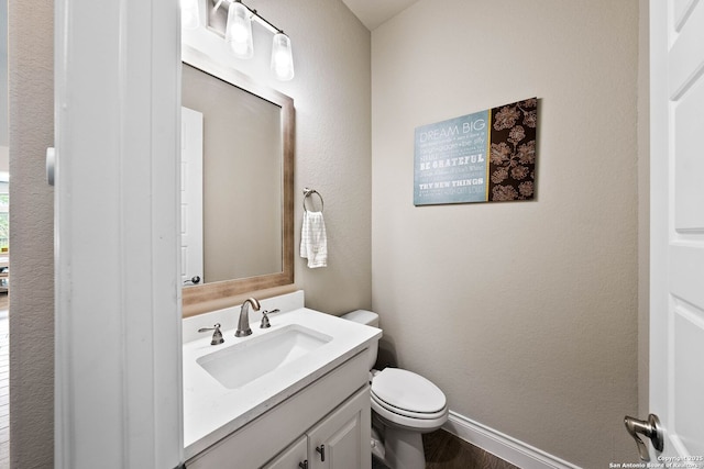 half bath featuring a textured wall, vanity, toilet, and baseboards