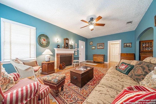 living room featuring arched walkways, light wood finished floors, visible vents, a brick fireplace, and ceiling fan