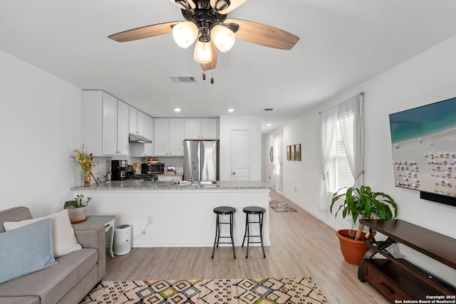 kitchen featuring freestanding refrigerator, a peninsula, light stone countertops, under cabinet range hood, and white cabinetry