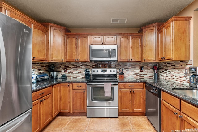 kitchen featuring appliances with stainless steel finishes, dark stone counters, brown cabinets, and visible vents