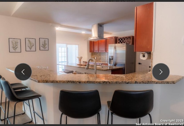 kitchen with light stone counters, island exhaust hood, stainless steel refrigerator with ice dispenser, and a peninsula