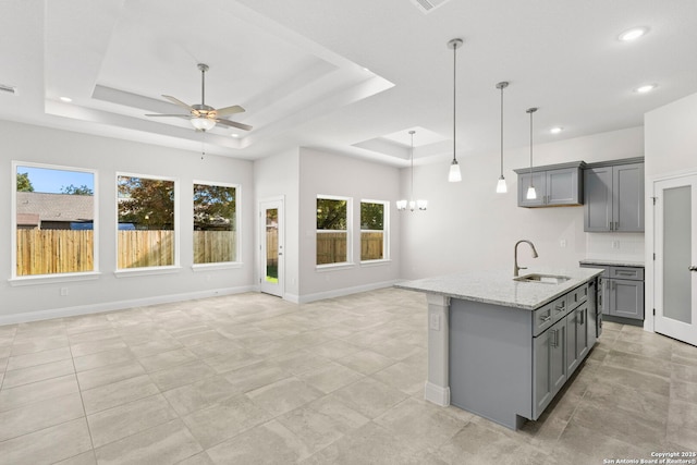 kitchen with light stone countertops, a kitchen island with sink, a tray ceiling, gray cabinetry, and a sink