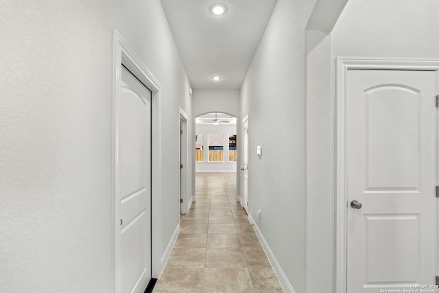 hall featuring arched walkways, a textured wall, light tile patterned flooring, and baseboards