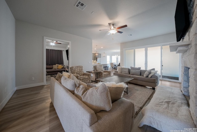 living room with visible vents, ceiling fan, a stone fireplace, wood finished floors, and baseboards