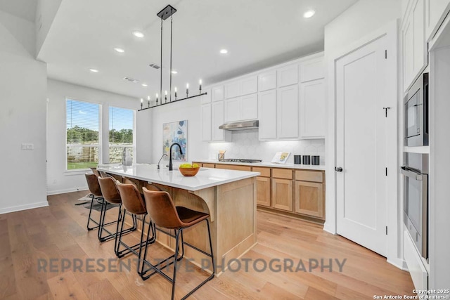 kitchen featuring white cabinets, an island with sink, decorative light fixtures, light countertops, and under cabinet range hood