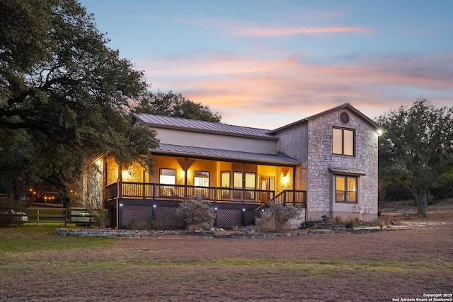 back of property at dusk with stone siding, metal roof, a standing seam roof, covered porch, and fence