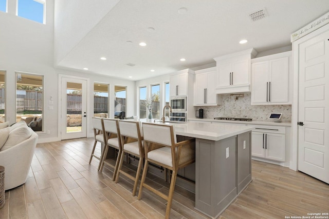 kitchen featuring visible vents, light wood finished floors, a kitchen island with sink, built in microwave, and tasteful backsplash