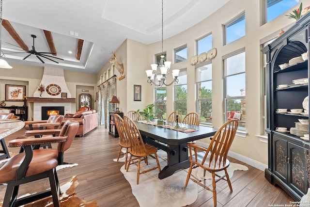 dining space with a large fireplace, ceiling fan with notable chandelier, wood finished floors, baseboards, and a tray ceiling