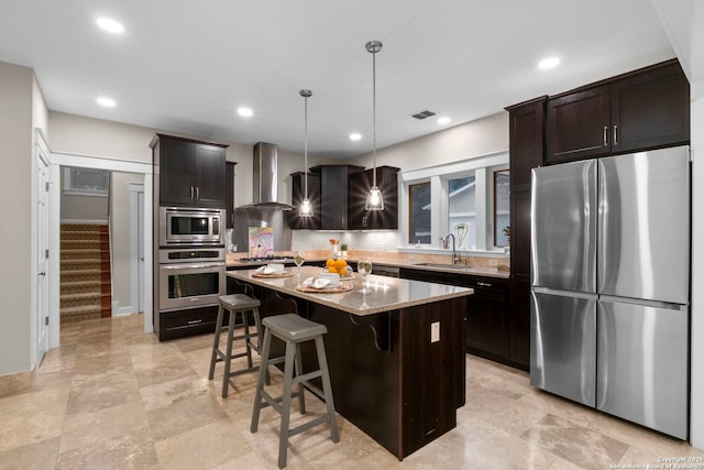 kitchen with light stone counters, stainless steel appliances, a kitchen island, hanging light fixtures, and wall chimney exhaust hood