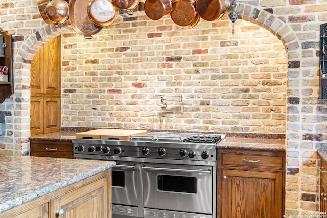 kitchen featuring brown cabinets, brick wall, dark stone counters, and range with two ovens