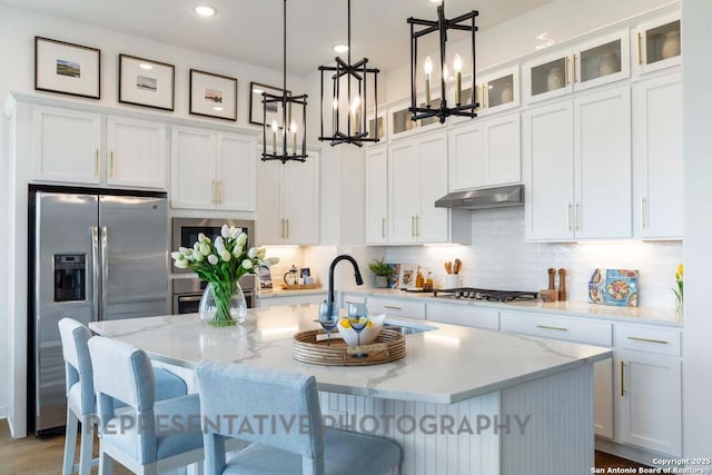 kitchen with under cabinet range hood, white cabinetry, appliances with stainless steel finishes, a center island with sink, and glass insert cabinets