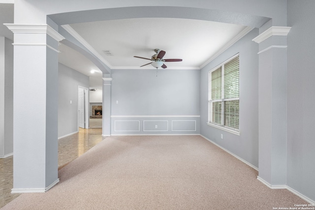 spare room featuring light carpet, a warm lit fireplace, decorative columns, a ceiling fan, and crown molding