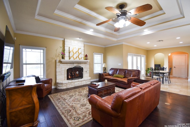 living room featuring arched walkways, a stone fireplace, a tray ceiling, and dark wood-type flooring