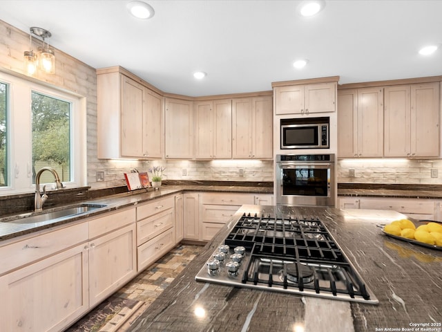 kitchen with appliances with stainless steel finishes, dark stone countertops, a sink, light brown cabinetry, and backsplash