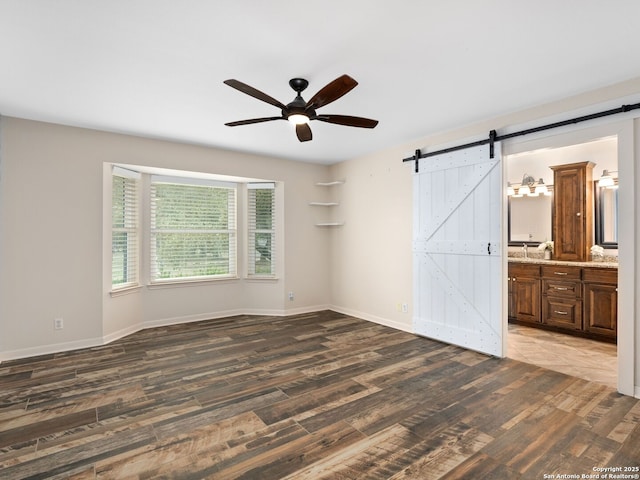 unfurnished bedroom featuring dark wood-style flooring, a barn door, ceiling fan, ensuite bath, and baseboards