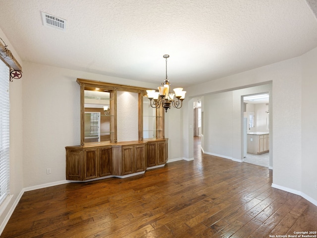 unfurnished dining area featuring dark wood-style floors, visible vents, a textured ceiling, a chandelier, and baseboards