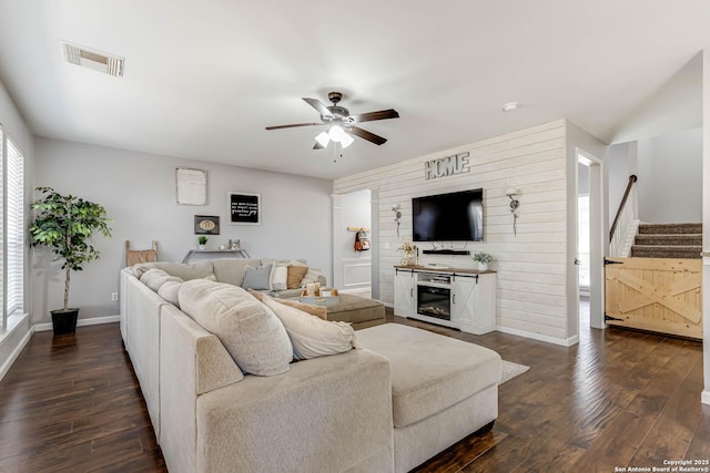 living room with dark wood-style floors, visible vents, a glass covered fireplace, ceiling fan, and stairs