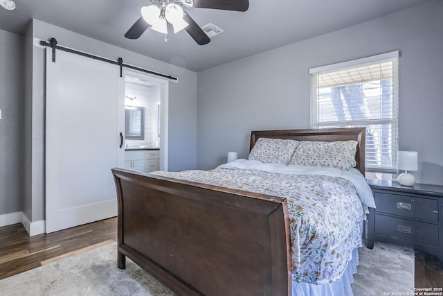 bedroom featuring dark wood-style floors, visible vents, a barn door, a ceiling fan, and ensuite bath