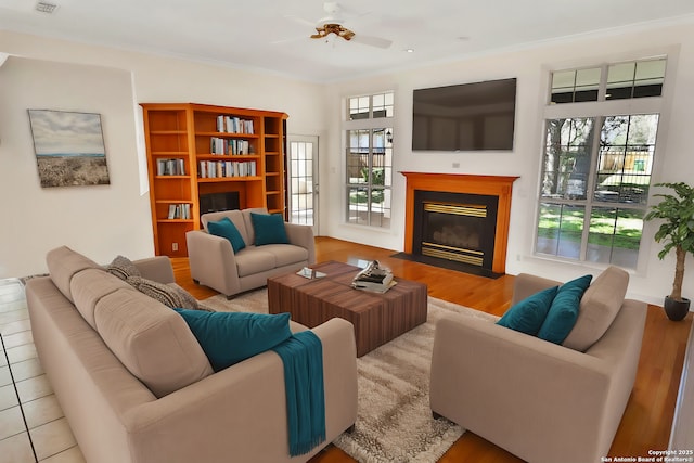 living room with light wood-style floors, a fireplace with flush hearth, ornamental molding, and ceiling fan