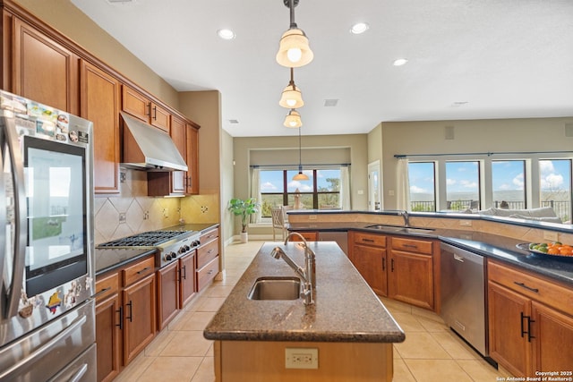 kitchen featuring pendant lighting, stainless steel appliances, a kitchen island with sink, a sink, and under cabinet range hood