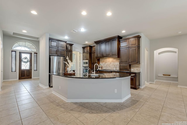 kitchen with arched walkways, light tile patterned floors, tasteful backsplash, a kitchen island with sink, and dark stone countertops