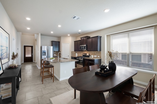 kitchen with visible vents, appliances with stainless steel finishes, dark brown cabinetry, an island with sink, and light stone countertops