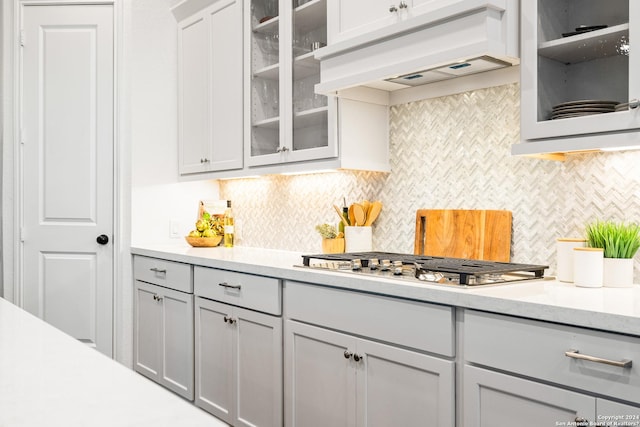 kitchen with stainless steel gas cooktop, light countertops, custom range hood, backsplash, and glass insert cabinets