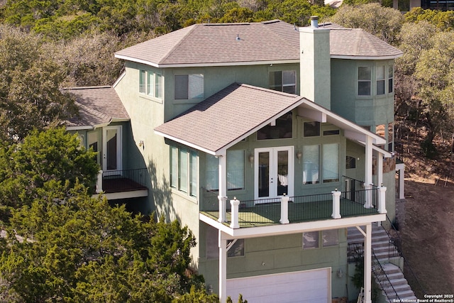 back of house featuring a balcony, a chimney, roof with shingles, french doors, and stucco siding