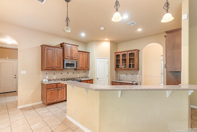 kitchen featuring visible vents, brown cabinets, stainless steel microwave, glass insert cabinets, and pendant lighting