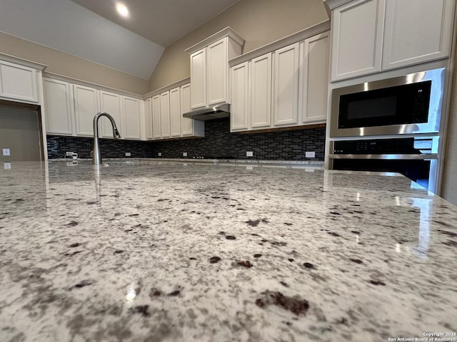 kitchen featuring light stone counters, under cabinet range hood, vaulted ceiling, appliances with stainless steel finishes, and backsplash