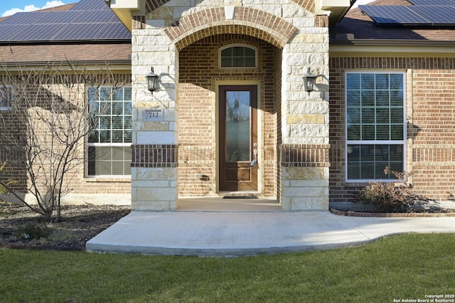 entrance to property featuring a shingled roof, stone siding, brick siding, and solar panels