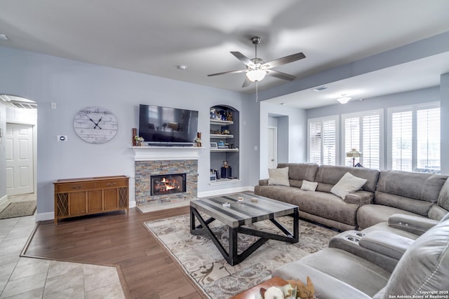 living room with a stone fireplace, built in shelves, wood finished floors, a ceiling fan, and baseboards