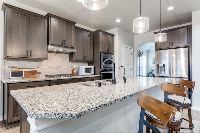 kitchen featuring arched walkways, under cabinet range hood, an island with sink, a kitchen bar, and decorative light fixtures