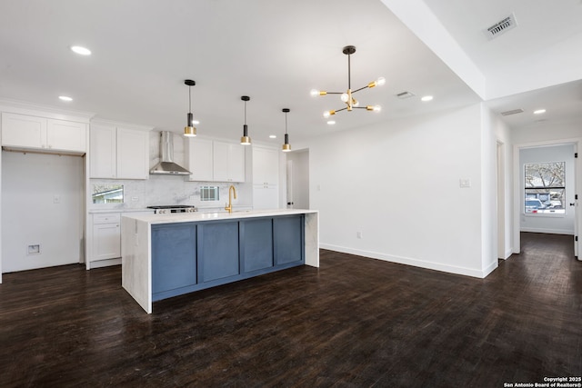 kitchen featuring wall chimney exhaust hood, a center island with sink, pendant lighting, and light countertops