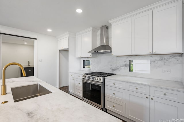 kitchen with a barn door, white cabinets, stainless steel gas stove, a sink, and wall chimney range hood