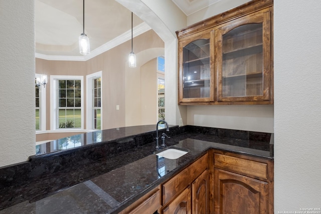 kitchen with hanging light fixtures, glass insert cabinets, ornamental molding, a sink, and dark stone countertops