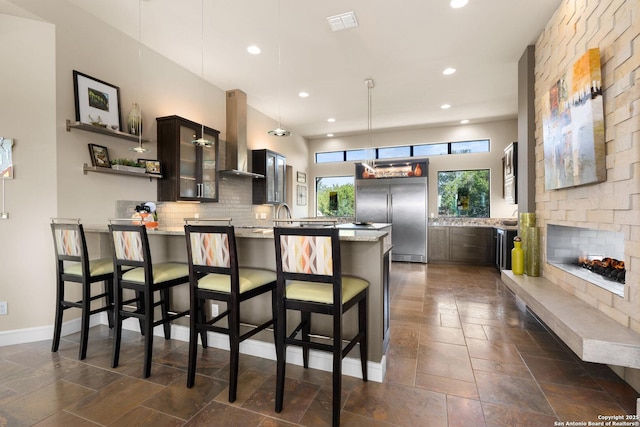 kitchen with glass insert cabinets, stainless steel built in fridge, a peninsula, and wall chimney exhaust hood