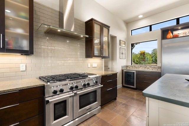 kitchen featuring dark brown cabinetry, tasteful backsplash, appliances with stainless steel finishes, glass insert cabinets, and wall chimney range hood