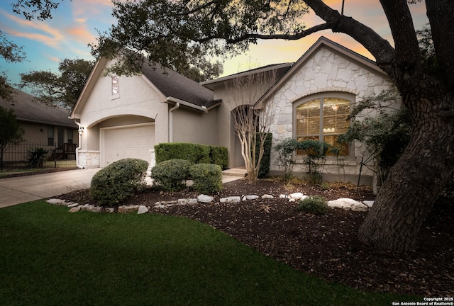 french country inspired facade with a garage, concrete driveway, stone siding, fence, and stucco siding