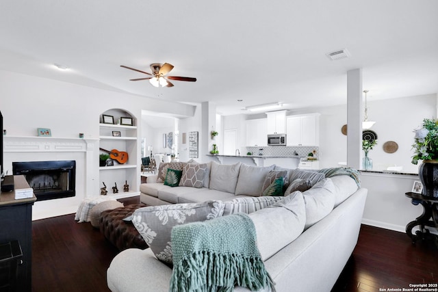 living room with built in features, dark wood-style flooring, a fireplace, visible vents, and a ceiling fan