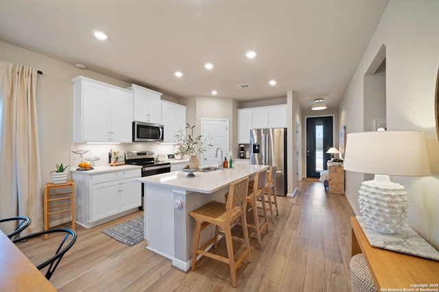 kitchen with light wood finished floors, stainless steel appliances, light countertops, white cabinetry, and an island with sink