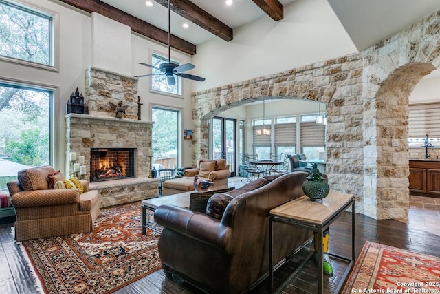 living room with arched walkways, a stone fireplace, dark wood-type flooring, a towering ceiling, and beam ceiling