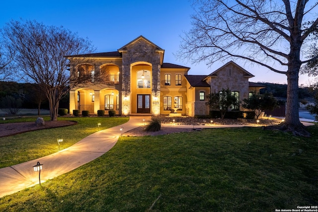 view of front facade featuring a balcony, stone siding, a front yard, and french doors