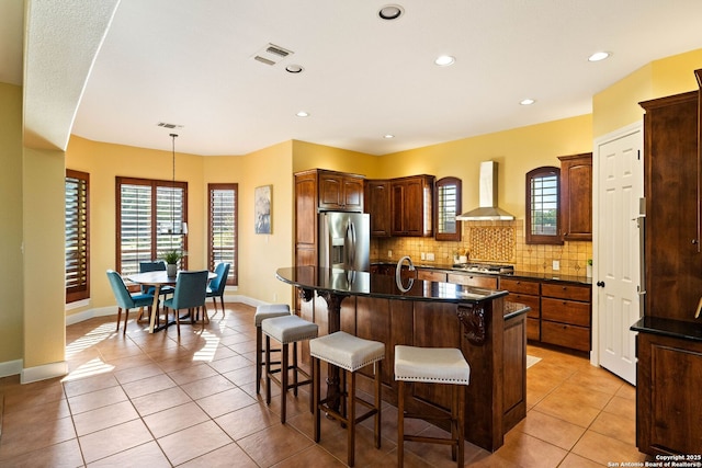 kitchen featuring a kitchen island with sink, stainless steel appliances, visible vents, wall chimney range hood, and dark countertops