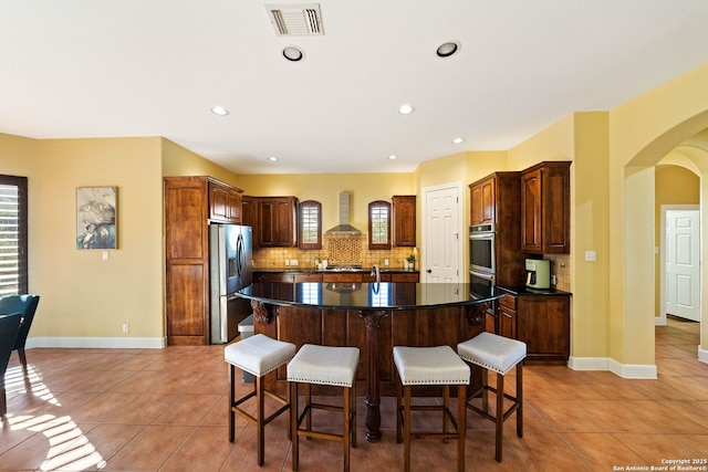 kitchen featuring arched walkways, a center island with sink, stainless steel appliances, dark countertops, and wall chimney range hood